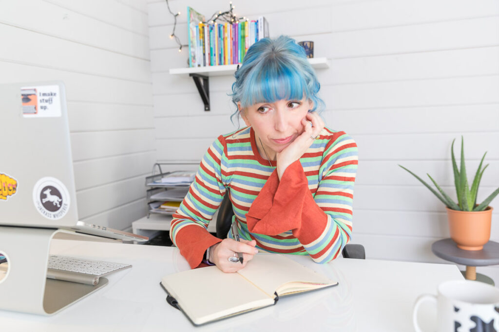 Vicky, with blue hair and an orange striped top, looks morose with notebook and pen in hand